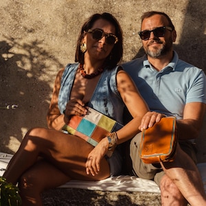 Può includere: Una coppia seduta su un muro di pietra, la donna tiene una pochette in pelle colorata e l'uomo tiene una borsa in pelle marrone.