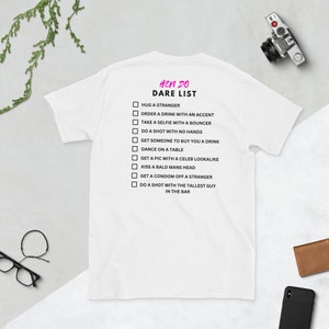 Puede incluir: Camiseta blanca con una lista de desafíos en negro para una despedida de soltera. El texto dice "Hen Do Dare List" e incluye desafíos como "Abrazar a un extraño", "Pedir una bebida con un acento" y "Tomar un trago con el hombre más alto del bar".