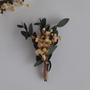 May include: A small bouquet of dried baby's breath flowers and eucalyptus leaves tied with twine. The bouquet is a light beige colour with green leaves.