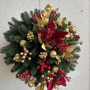 May include: A round Christmas wreath with evergreen branches, red berries, gold ornaments, and burgundy poinsettias. The wreath is suspended from a chain and is against a white textured wall. The wreath is a festive holiday decoration.