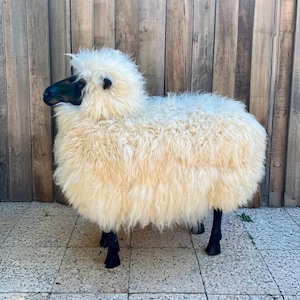 May include: A whimsical sheep-shaped stool with a fluffy, cream-colored wool body and black legs and face. The decorative stool is placed on a tiled surface in front of a wooden wall. A potted plant and a chair are in the background.