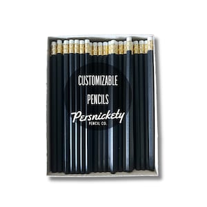Puede incluir: Una caja de 24 lápices hexagonales negros con gomas blancas y detalles dorados. La caja tiene una etiqueta en blanco y negro que dice "CUSTOMIZABLE PENCILS Persnickety PENCIL CO."