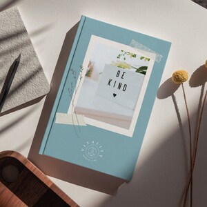 May include: A blue journal with a white photo of a sign that says "Be Kind" with a small heart. The journal has a logo that says "Vitality Whole Life" with a stylized image of a person meditating.