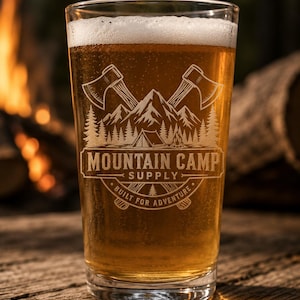 May include: A clear glass pint filled with golden beer, featuring an etched design of crossed axes, a mountain range, a tent, and a campfire. The text reads MOUNTAIN CAMP SUPPLY and BUILT FOR ADVENTURE, centered within a circular emblem.