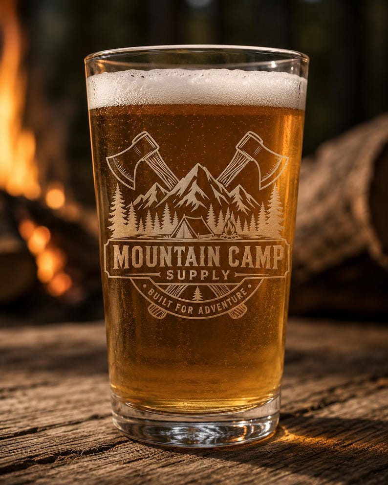 May include: A clear glass pint filled with golden beer, featuring an etched design of crossed axes, a mountain range, a tent, and a campfire. The text reads MOUNTAIN CAMP SUPPLY and BUILT FOR ADVENTURE, centered within a circular emblem.