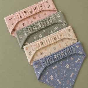 May include: Four dog bandanas in pink, green, beige, and blue. Each bandana features a repeating pattern of paw prints, bones, and botanical elements. The blue bandana has moons and stars. The bandanas are folded and arranged on a green surface.