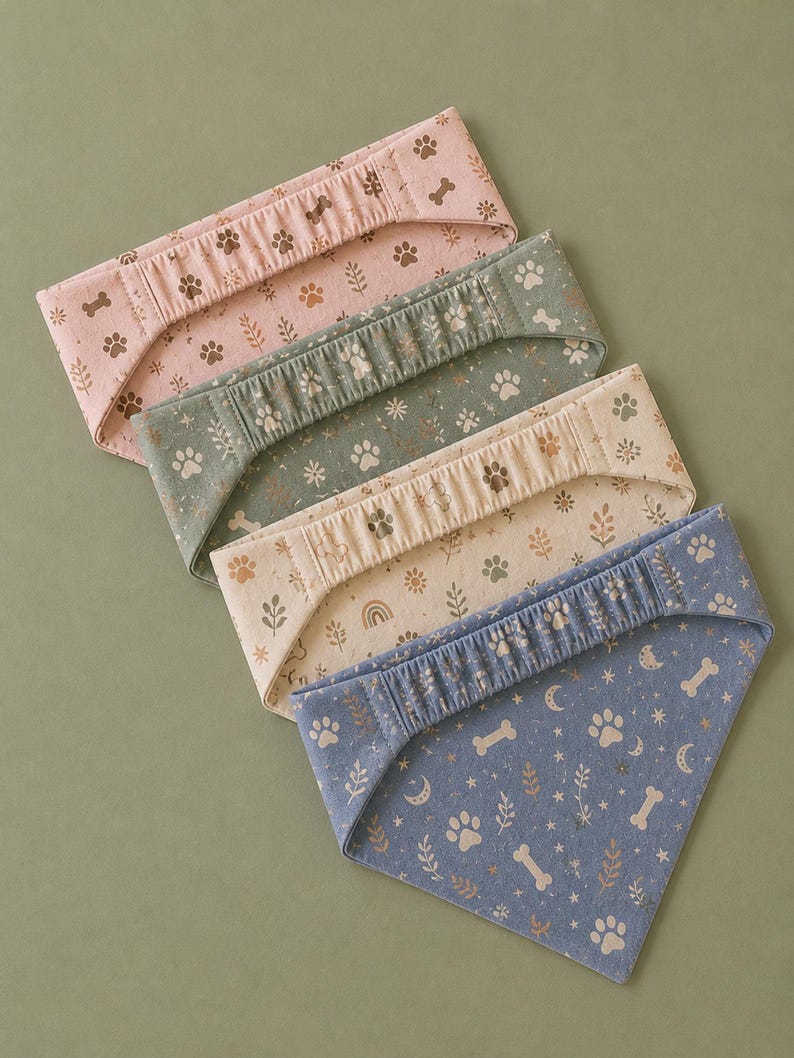 May include: Four dog bandanas in pink, green, beige, and blue. Each bandana features a repeating pattern of paw prints, bones, and botanical elements. The blue bandana has moons and stars. The bandanas are folded and arranged on a green surface.
