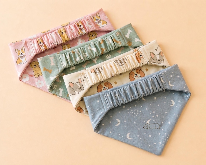 May include: Four dog bandanas in pink, green, cream, and blue. Each bandana features a different pattern of dogs, bones, and stars. The bandanas have an elasticated top and a triangular shape.