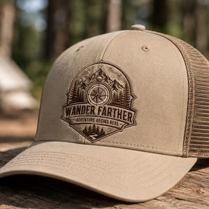 May include: A tan trucker hat featuring a brown embroidered patch on the front. The patch displays a mountain range, a compass, pine trees, and a winding path with the text WANDER FARTHER and ADVENTURE BEGINS HERE in a circular design.