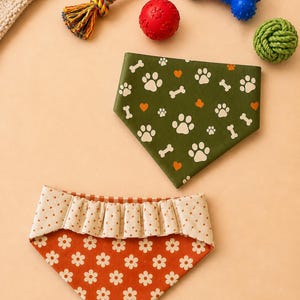 May include: Two dog bandanas with different patterns. One is green with white paw prints, orange hearts, and bone shapes. The other is orange with white flower shapes and a ruffled top. Also, dog toys including a rope toy, a red ball, a blue bone, and a green rope ball.