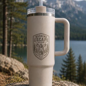 May include: A matte beige insulated tumbler with a handle, a clear lid, and a straw. The front features a dark brown graphic of a lantern, pine trees, and mountains with the text "WEEKEND ESCAPE CLUB" and "LEAVE THE CITY BEHIND" in a shield.
