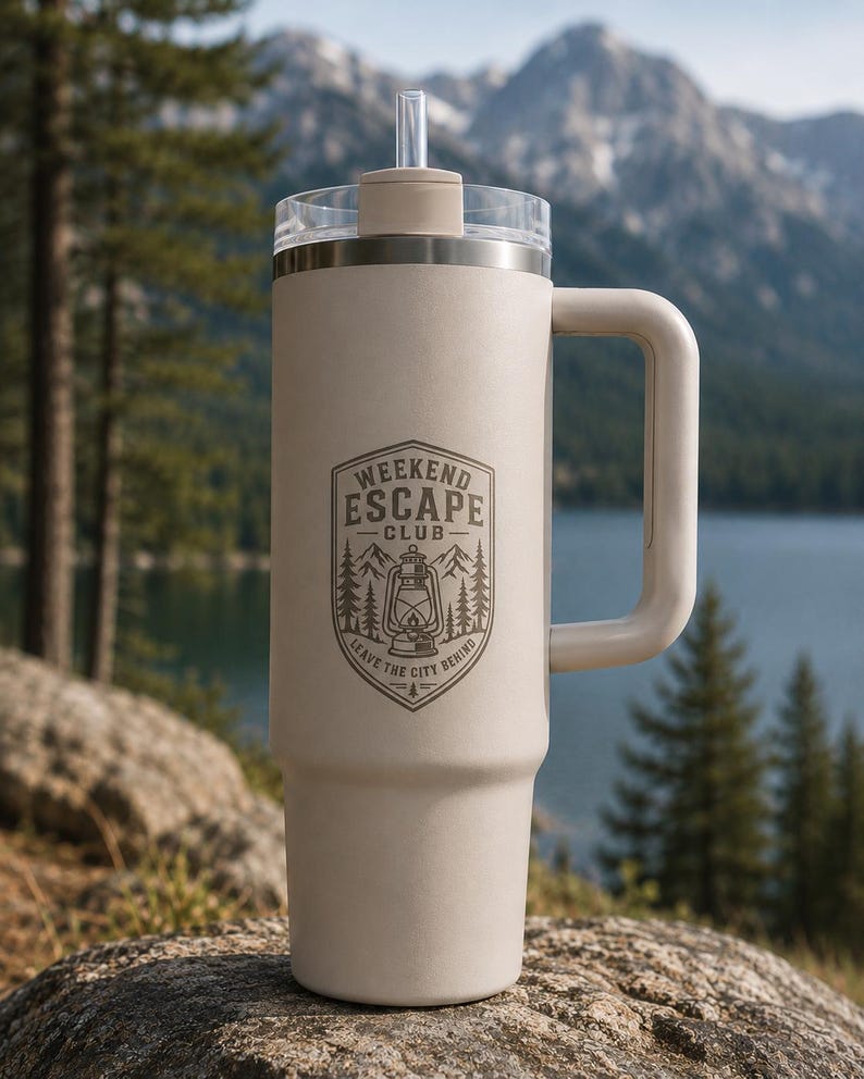 May include: A matte beige insulated tumbler with a handle, a clear lid, and a straw. The front features a dark brown graphic of a lantern, pine trees, and mountains with the text "WEEKEND ESCAPE CLUB" and "LEAVE THE CITY BEHIND" in a shield.