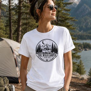 May include: A white short-sleeved t-shirt featuring a circular black graphic print. The design depicts a camper van, pine trees, mountains, and a crescent moon, with the text THE WILD CAMPER and ROAM FREE centered below the illustration.