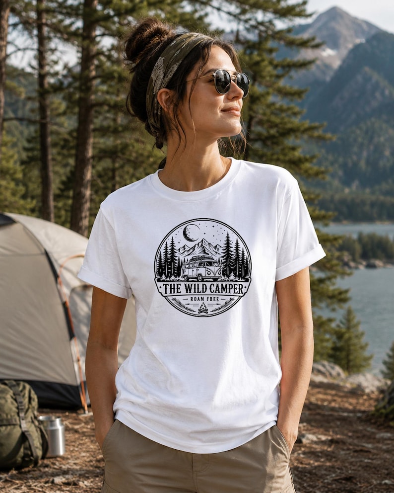 May include: A white short-sleeved t-shirt featuring a circular black graphic print. The design depicts a camper van, pine trees, mountains, and a crescent moon, with the text THE WILD CAMPER and ROAM FREE centered below the illustration.