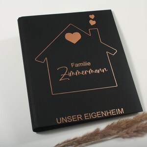 Könnte beinhalten: Ein schwarzes Buch mit einem kupferfarbenen Hausdesign, Herz und Text. Der Text enthält "Familie Zimmermann" und "UNSER EIGENHEIM". Das Buch liegt auf einer weißen Oberfläche mit getrocknetem Ziergras.