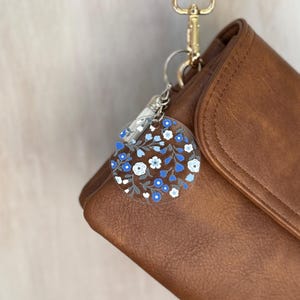 May include: A clear, round keychain with a floral design of blue and white flowers and gray leaves. The keychain is attached to a brown leather bag with a gold-colored clasp and strap.