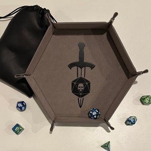 May include: A gray, hexagonal dice tray with a black sword and skull design, a black drawstring bag, and several multi-sided dice. The tray has metal clasps at each corner. The dice are blue, green, and clear.
