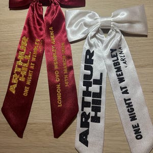 以下が含まれることがあります： 2つのサテンリボン。1つは、金色の文字で「ARTHUR HILL ONE NIGHT AT WEMBLEY ARENA」と「LONDON, OVO WEMBLEY ARENA 19TH MARCH」と書かれたバーガンディ色です。もう1つは、黒い文字で「ARTHUR HILL ONE NIGHT AT WEMBLEY ARENA」と書かれた白です。