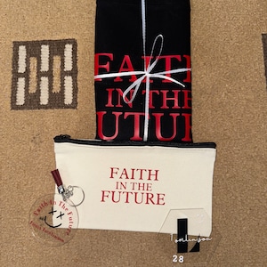 May include: A black tote bag with white ribbon and the text "FAITH IN THE FUTURE" in red. A white canvas pouch with the same text in red. A clear acrylic keychain with the text "Faith in the Future" and a smiley face.