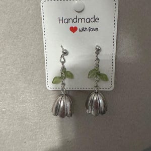 May include: A pair of handmade silver-colored drop earrings. Each earring features a small silver ball, a chain, a green leaf, and a silver flower-shaped charm. The earrings are displayed on a white card that reads "Handmade with love."