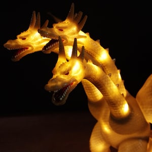 May include: A three-headed dragon figurine made of a translucent, off-white material that glows with an internal warm yellow light. Each head features sharp horns, visible scales, and an open mouth with red accents, illuminated from within by LEDs.