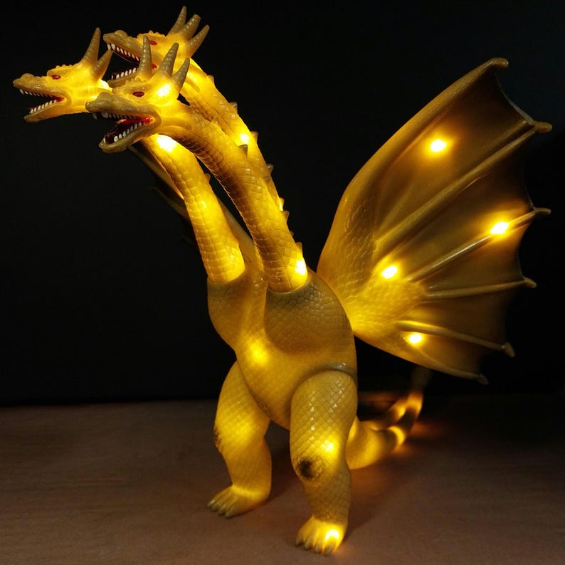 May include: A yellow three-headed dragon figurine with textured scales and large wings. The creature is illuminated from within by multiple warm yellow lights visible through the translucent material, highlighting the heads, necks, and wings.