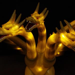 May include: A three-headed golden dragon figurine with illuminated yellow LED lights throughout its body. Each head features sharp horns, red eyes, and an open mouth with visible white teeth. The creature has extended wings and a textured scale pattern.