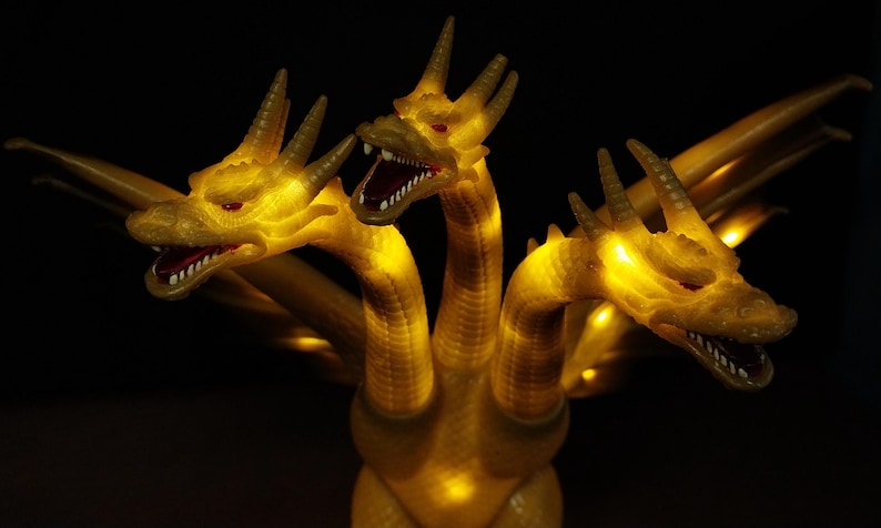 May include: A three-headed golden dragon figurine with illuminated yellow LED lights throughout its body. Each head features sharp horns, red eyes, and an open mouth with visible white teeth. The creature has extended wings and a textured scale pattern.
