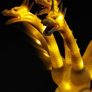 May include: A three-headed dragon figurine with a translucent yellow body that glows from internal lighting. Each head features sharp teeth, red eyes, and textured scales. The creature has long necks and wings visible against a dark background.