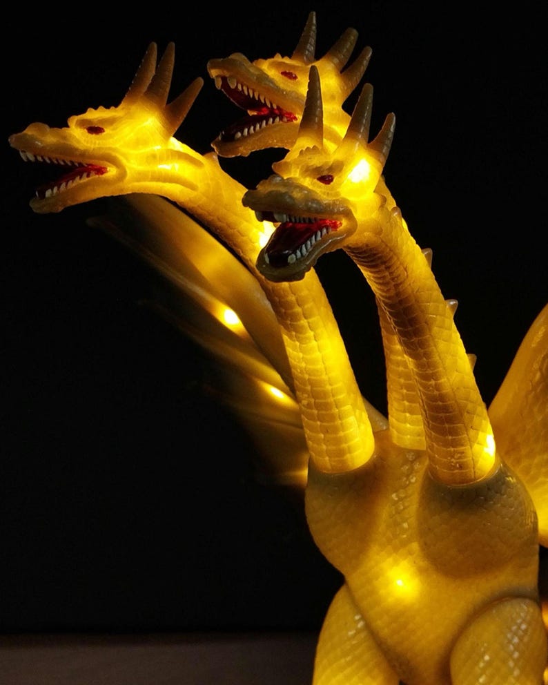 May include: A three-headed dragon figurine with a translucent yellow body that glows from internal lighting. Each head features sharp teeth, red eyes, and textured scales. The creature has long necks and wings visible against a dark background.
