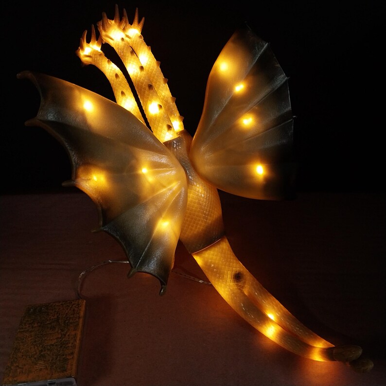May include: A three-headed dragon lamp with translucent off-white skin and textured scales. The creature features large wings and a long tail, all illuminated from within by warm yellow LED lights. A rectangular battery pack is connected by a wire.