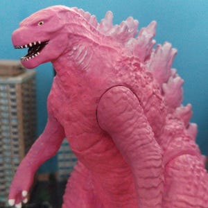 Könnte beinhalten: Eine pinkfarbene Godzilla-Actionfigur mit detaillierter, strukturierter Haut und Rückenflossen. Die Figur hat ein weit geöffnetes Maul mit sichtbaren Zähnen und gelben Augen. Der Hintergrund ist hellblau.