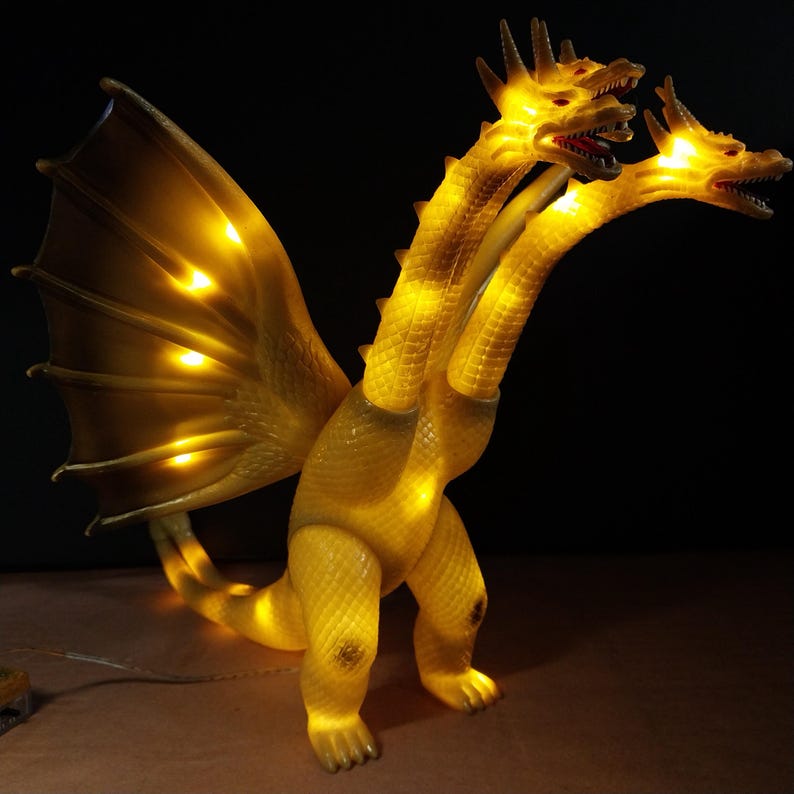 May include: A three-headed yellow dragon lamp with a textured, scaly body and large wings. The creature is illuminated from within by warm yellow lights, highlighting its three open mouths, sharp teeth, and detailed wing structure against a dark background.