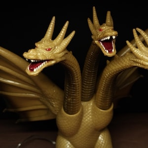 May include: A three-headed golden dragon figurine with textured scales, large wings, and red eyes. Each head features an open mouth with visible white teeth and a dark red interior. The creature has a long neck for each head and a scaly body.