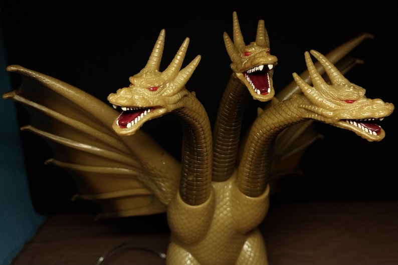 May include: A three-headed golden dragon figurine with textured scales, large wings, and red eyes. Each head features an open mouth with visible white teeth and a dark red interior. The creature has a long neck for each head and a scaly body.