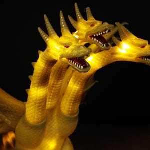 May include: A three-headed golden dragon figurine featuring illuminated yellow eyes and mouths. The creature has textured scales, sharp horns on each head, and visible wings. The internal lighting creates a glowing effect throughout the body.