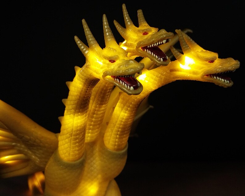 May include: A three-headed golden dragon figurine featuring illuminated yellow eyes and mouths. The creature has textured scales, sharp horns on each head, and visible wings. The internal lighting creates a glowing effect throughout the body.