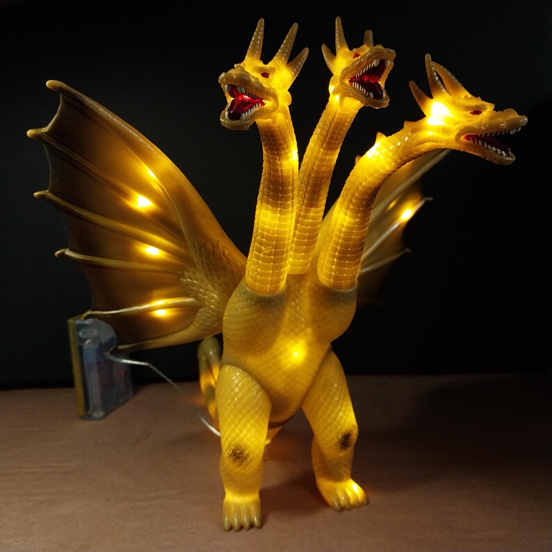 May include: A three-headed golden dragon figure with illuminated yellow LED lights throughout its body and wings. The creature features textured scales, open mouths with red interiors, and a wired battery pack connected to the base of the wings.