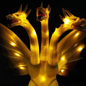 May include: A three-headed dragon figurine with translucent yellow skin and integrated internal LED lights that illuminate the necks and wings. Each head features open jaws with visible teeth and red eyes, set against a solid black background.