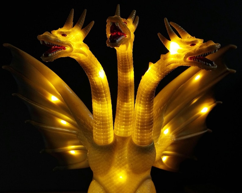 May include: A three-headed dragon figurine with translucent yellow skin and integrated internal LED lights that illuminate the necks and wings. Each head features open jaws with visible teeth and red eyes, set against a solid black background.