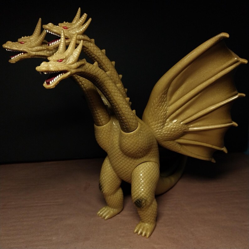 May include: A gold-coloured three-headed dragon figurine with textured scales, red eyes, and large wings. The creature stands on two legs with a long tail visible behind it. Each of the three heads features sharp horns and an open mouth with visible teeth.