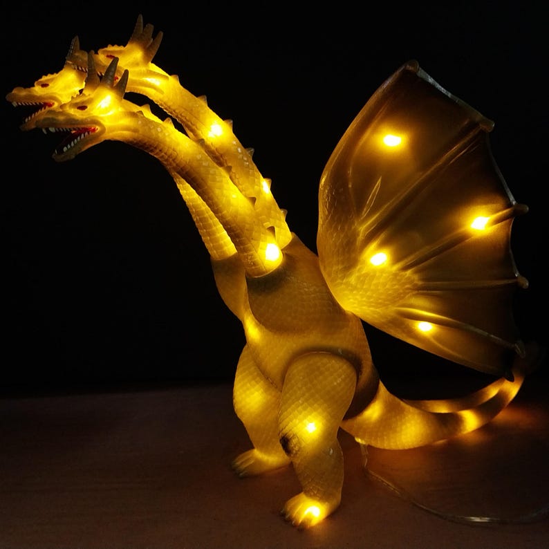 May include: A three-headed dragon figurine made of translucent yellow material, illuminated from within by multiple bright LED lights. The creature features textured scales, open mouths with visible teeth, and large wings, all glowing with a warm golden light against a dark background.