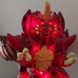 May include: A red, illuminated monster figurine with a menacing expression. The figure has a large head with a horn, sharp teeth, and glowing yellow eyes. The body features a textured design with a yellow leaf-like detail.