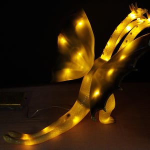 May include: A three-headed dragon lamp with a translucent, textured body that glows with internal yellow light. The creature features large wings, a long tail, and three distinct heads, all illuminated from within, creating a warm, bright effect.