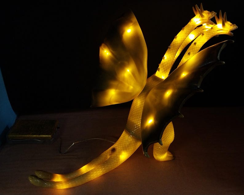 May include: A three-headed dragon lamp with a translucent, textured body that glows with internal yellow light. The creature features large wings, a long tail, and three distinct heads, all illuminated from within, creating a warm, bright effect.