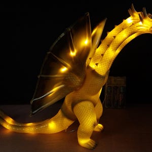 May include: A three-headed dragon figurine with a textured, translucent yellow body that glows from internal lighting. The creature features large, pointed wings, a long tail, and multiple necks extending upward, all illuminated in a warm golden hue.