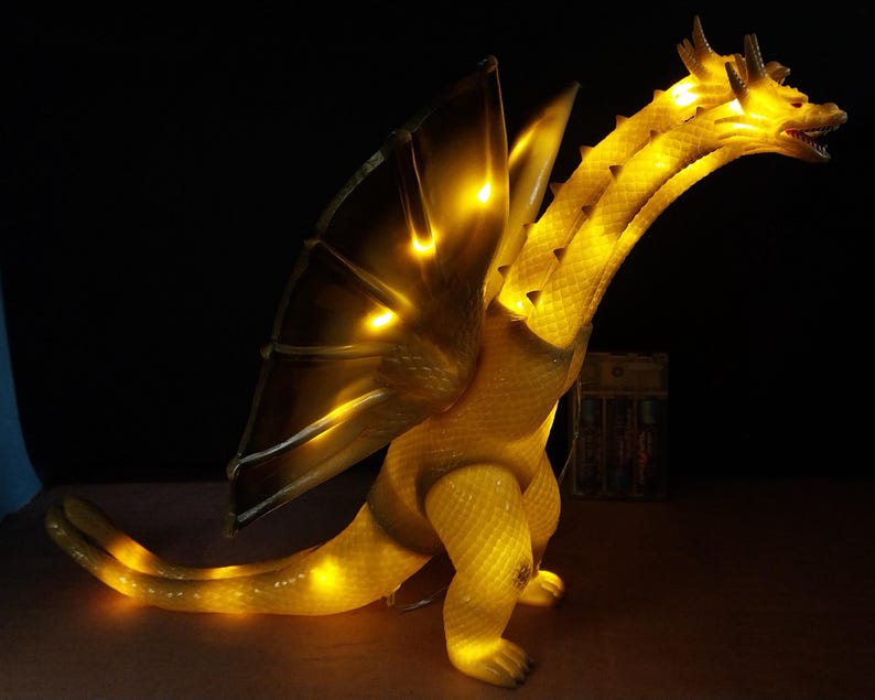 May include: A three-headed dragon figurine with a textured, translucent yellow body that glows from internal lighting. The creature features large, pointed wings, a long tail, and multiple necks extending upward, all illuminated in a warm golden hue.