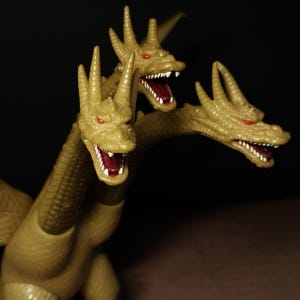 May include: A three-headed dragon figurine with textured tan skin, sharp horns on each head, and small red eyes. Each of the three open mouths reveals white teeth and a dark red interior, showcasing a detailed reptilian design on each neck.
