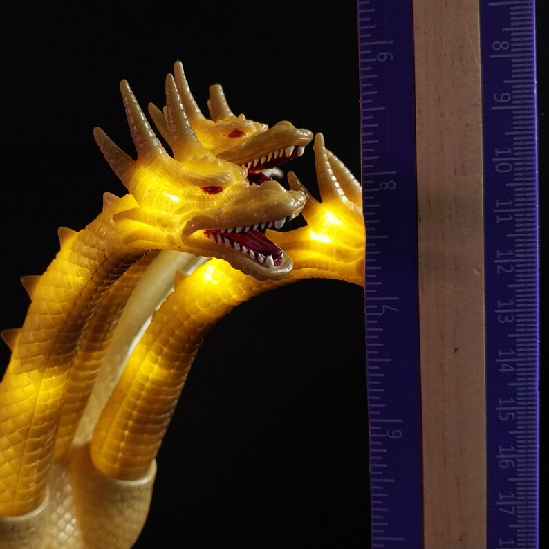 May include: A three-headed yellow dragon figurine with glowing internal lights, featuring textured scales and open mouths with visible teeth. A wooden ruler is positioned vertically next to the dragon, showing a height of approximately 18 cm.