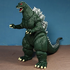 May include: A dark green Godzilla action figure with a textured, scaly body, sharp white teeth, and red eyes. The creature features prominent silver-colored dorsal fins running down its back and a long tail, with light-colored claws on its feet.
